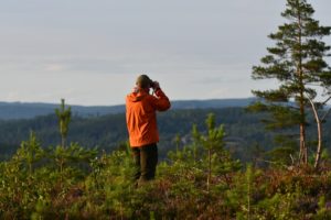 Person i orange jacka står med kikare på en höjd i naturen